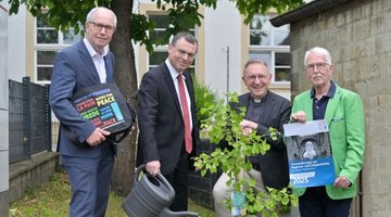 Das Foto zeigt einen jungen Ginkgo-Baum am Forum St. Liborius, der von (v.l.n.r.) Landrat a. D. Manfred Müller, Pfarrer Dr. Eckhard Düker (Evangelischer Kirchenkreis Paderborn), Dechant und Domkapitular Benedikt Fischer (Erzbistum) und Hermann-Josef Bentler als Kreisvorstandsmitglied des Volksbundes) gepflanzt wurde. Pfarrer Dr. Eckhard Düker gießt den Baum noch, während Dechant und Domkapitular Benedikt Fischer den Spaten der Baumpflanzaktion noch in den Händen hält.